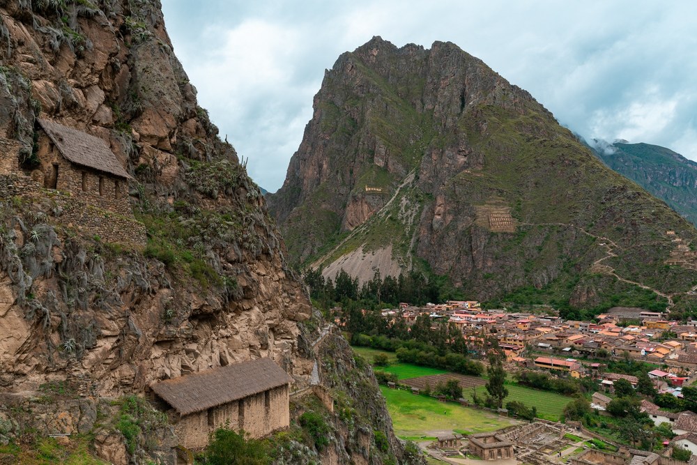 Stone walls and a hillside building overlook Ollantaytambo, Peru, beneath a steep mountain.
